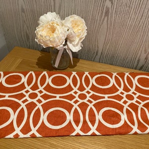 May include: A terracotta-coloured table runner featuring a white geometric design of overlapping circles. A small glass vase holds a bouquet of cream-coloured artificial flowers, tied with a pink ribbon. The runner is on a wooden table.