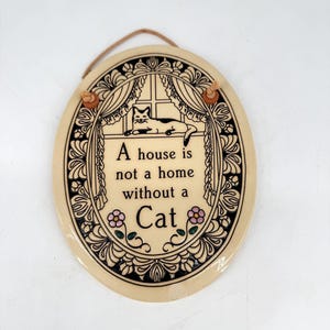 May include: Cream-coloured, oval ceramic plaque with a black decorative border. The plaque features a cat in a window, with the text "A house is not a home without a Cat". Two pink flowers are at the bottom.