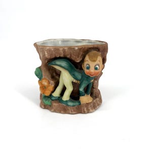 Puede incluir: Una maceta de cerámica vintage con forma de tronco de árbol con una figura caprichosa. La figura está vestida con un atuendo verde y crema, con una cara sonriente y ojos azules. La maceta tiene un exterior marrón y un interior blanco.