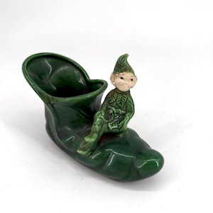 Peut inclure: Figurine vintage en céramique représentant une jardinière en forme de chaussure verte avec une figurine d'elfe assis. L'elfe est vêtu de vert avec un chapeau pointu. La jardinière et l'elfe ont une finition brillante, sur fond blanc.