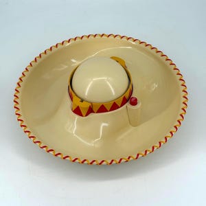 Pode incluir: Um cinzeiro em forma de chapéu mexicano creme, com uma cúpula central e uma guarnição vermelha, amarela e creme. A aba é adornada com costuras vermelhas e amarelas. Um pequeno botão vermelho está na lateral.