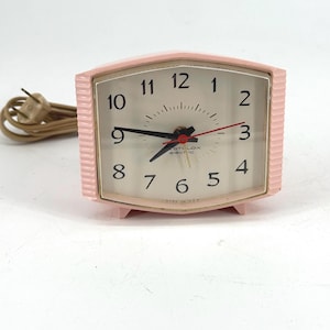 May include: A retro pink electric clock with a rectangular face and a ribbed frame. The clock face features black numerals, black hands, and a red second hand. The clock is labelled "STCLOX electric" and has a beige power cord.