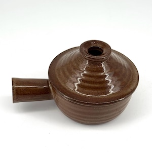 Könnte beinhalten: Eine braune Keramik-Auflaufform mit Deckel und Griff. Die Form hat einen runden Körper und einen Deckel mit einer kleinen Öffnung oben. Der Griff ist rechteckig und erstreckt sich von der Seite der Form.