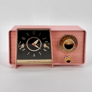 Puede incluir: Un radio despertador vintage rosa pálido General Electric con una esfera de reloj negra y un dial dorado. La radio tiene una función de reposo y alarma.