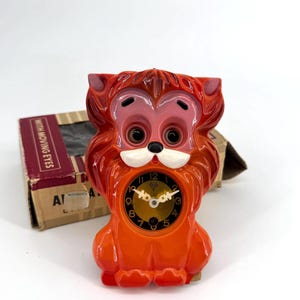 May include: A vintage orange lion-shaped clock with a black clock face and gold hands. The clock has a cartoonish design with large eyes and a white muzzle. The clock is next to a cardboard box with the text "WITH MOVING EYES".