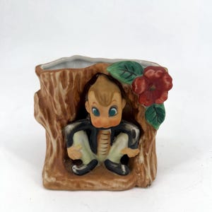 Op de afbeelding: Vintage keramische plantenbak in de vorm van een boomstronk, met een zittend stripfiguur met blauwe ogen en een zwart jasje. Een rode bloem en groene bladeren sieren de zijkant. De plantenbak heeft een bruin en beige kleurenschema.