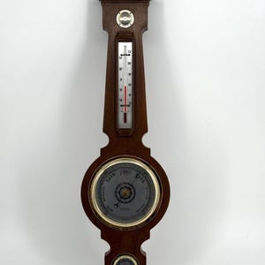 Puede incluir: Un barómetro de madera vintage con termómetro y un indicador circular. El termómetro muestra una temperatura de aproximadamente 21 grados Celsius. El barómetro tiene un acabado marrón oscuro y un borde dorado.