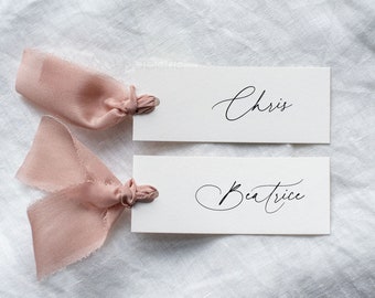 Könnte beinhalten: Zwei weiße Platzkarte mit kalligrafischen Namen, Chris und Beatrice, gebunden mit Schleifen aus rosafarbenem Band.