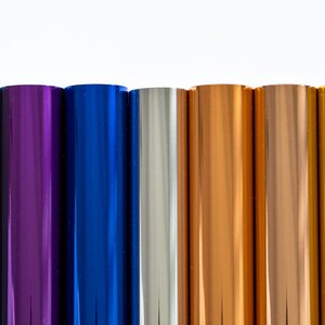 May include: A selection of metallic foil rolls in various colours, including green, purple, blue, silver, copper, and gold. The rolls are displayed vertically against a white backdrop, highlighting their shiny surfaces.