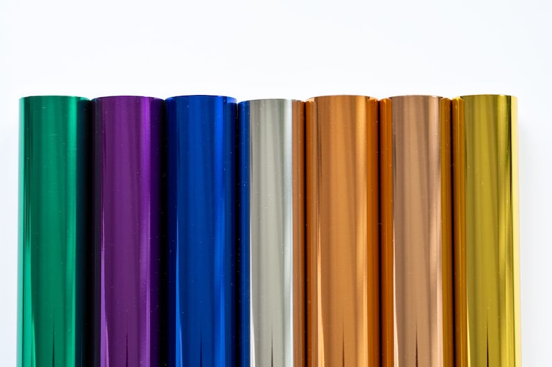May include: A selection of metallic foil rolls in various colours, including green, purple, blue, silver, copper, and gold. The rolls are displayed vertically against a white backdrop, highlighting their shiny surfaces.