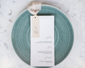 May include: A white wedding menu card with a beige tag that says "Bride" on it. The menu includes a starter, main course, and dessert. The card is on a teal plate with a white ribbon.