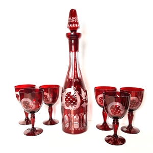 May include: A ruby red glass decanter with a stopper and six matching wine glasses. The set features etched floral and geometric designs. The decanter is tall and slender, and the glasses have a classic wine glass shape.