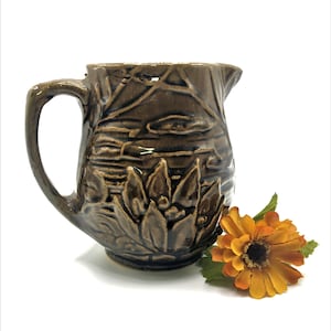 May include: A brown ceramic pitcher with a floral design. The pitcher has a wide mouth and a handle. The design features a stylized lily pad and leaves.