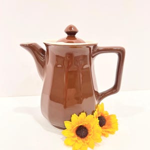 May include: A brown ceramic coffee pot with a lid and handle. The pot has a glossy finish and a white interior rim. Two yellow sunflowers with brown centers are placed near the base of the pot.