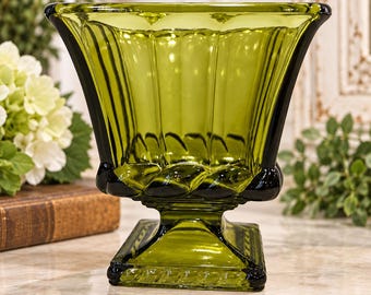 May include: A translucent olive green glass vase featuring a wide, flared opening with vertical fluted panels along the sides. The vessel rests on a square pedestal base with a decorative rope-like texture around the bottom edge.