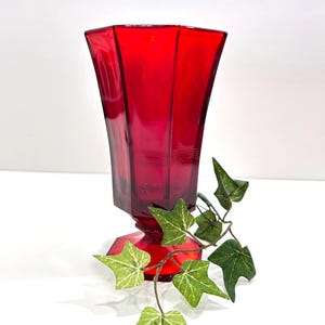 May include: A ruby red glass vase with a geometric design. The vase has a flared top and a pedestal base. Green ivy with white veins is draped around the vase.