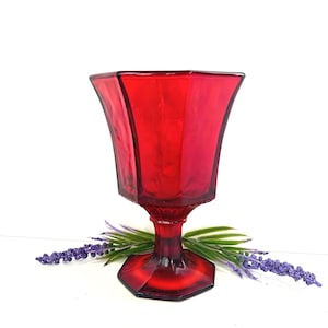 May include: A ruby red, vintage-style glass goblet with a hexagonal base and bowl. The goblet has a flared, faceted design and a short stem. The glass is a deep, transparent red. The goblet is decorated with purple and green faux flowers.