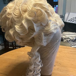 Może przedstawiać: Jasna blond peruka z obszernymi, ciasno skręconymi lokami. Peruka jest stylizowana w klasycznym, historycznym stylu, z lokami kaskadowo opadającymi na plecy. Umieszczona na głowie manekina.