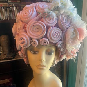 May include: A mannequin head wearing a sculpted wig made of numerous light pink foam rolls arranged in a dense, circular pattern. The wig is adorned with a cluster of white and pale pink artificial flowers on the right side. A white tag reads Wigs By Byron.