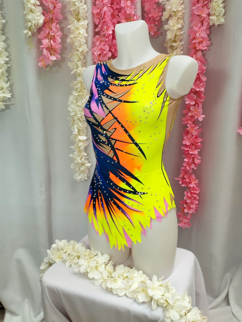 May include: A yellow, pink, orange, and blue leotard with a geometric design and rhinestones. The leotard has a scoop neck and a sleeveless design.