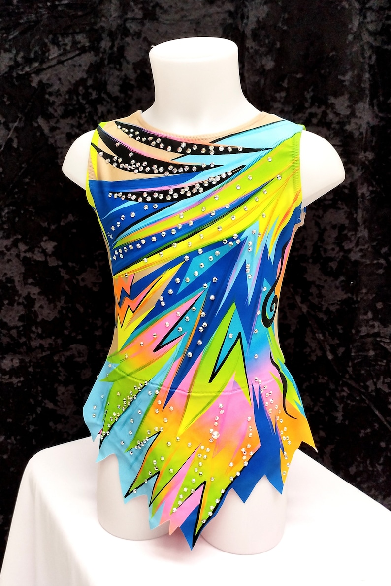 May include: A colorful leotard for gymnastics or dance, displayed on a mannequin. The sleeveless garment features a vibrant design with blue, green, yellow, and pink geometric patterns, accented with sparkling rhinestones. The leotard has a high neckline and a unique cut.