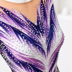 Op de afbeelding: Een mouwloze turnpakje met een doorschijnende halslijn, versierd met een levendig paars en wit veervormig ontwerp. Het turnpakje is versierd met talrijke sprankelende strass-steentjes. Het kledingstuk is ontworpen voor gymnastiek of dans.