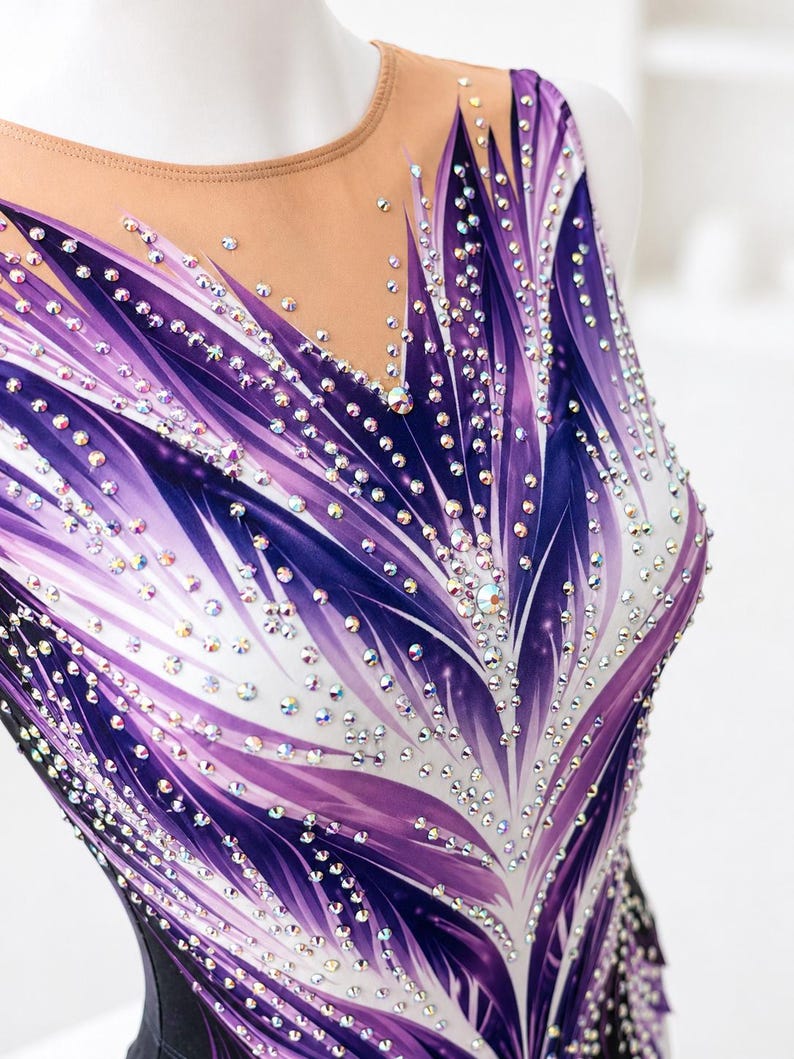 Op de afbeelding: Een mouwloze turnpakje met een doorschijnende halslijn, versierd met een levendig paars en wit veervormig ontwerp. Het turnpakje is versierd met talrijke sprankelende strass-steentjes. Het kledingstuk is ontworpen voor gymnastiek of dans.
