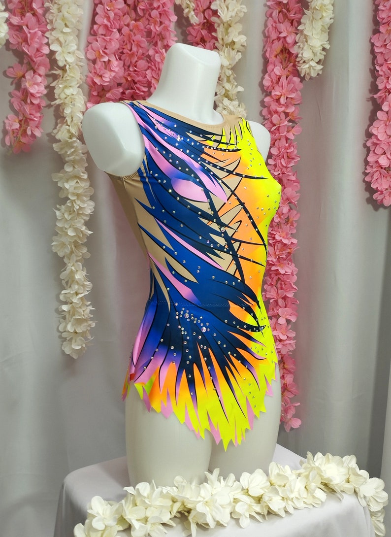 May include: A colorful leotard with a blue, pink, yellow, and orange abstract design. The leotard has a scoop neck and a sleeveless design. It is adorned with rhinestones.
