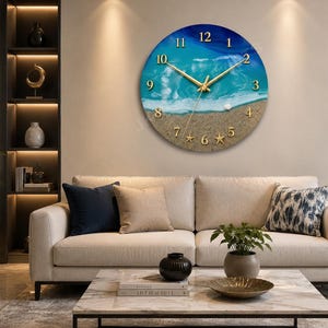 May include: A round wall clock featuring a beach scene with blue ocean waves, white sea foam, and sandy shore. The clock face includes gold-colored numbers from 1 to 12, gold-colored clock hands, and small starfish accents along the bottom edge.
