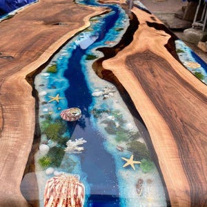 Puede incluir: Mesa de madera artesanal con un río de resina azul en el centro. El río de resina está decorado con conchas marinas, estrellas de mar y coral, creando un tema costero. Los bordes de madera de la mesa son marrones.
