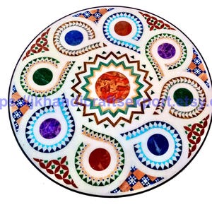 Op de afbeelding: Ronde witte marmeren tafelblad met een ingewikkeld ingelegd ontwerp. Het ontwerp heeft een centraal zonnesterpatroon in tinten oranje, bruin en groen, omgeven door kleurrijke wervelingen en geometrische vormen. Het tafelblad heeft een zwarte rand.