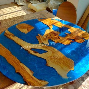 Puede incluir: Mesa redonda con una vibrante superficie de resina epoxi azul, con incrustaciones de elementos de madera natural. La madera es de color marrón claro y tiene un patrón de vetas único. El texto "JKHANDICRAFTS" es visible.