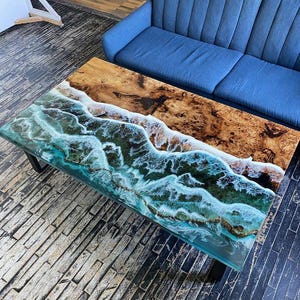 Puede incluir: Mesa de centro rectangular con un diseño único. La parte superior presenta un patrón de vetas de madera en una mitad y una resina transparente con un diseño de olas en la otra. La mesa está soportada por patas de metal negro.