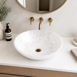 May include: A round, white vessel sink with a shimmering, iridescent mother-of-pearl finish. The basin features a central drain with a metallic gold-toned finish, matching the wall-mounted faucet and handles positioned above the white countertop.