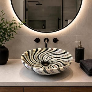 Puede incluir: Un lavabo de sobreponer redondo con un patrón de remolinos de segmentos de piedra en colores negro y crema alternados. El cuenco tiene un acabado liso y pulido con un desagüe central negro. Está montado sobre una encimera clara con un grifo negro.