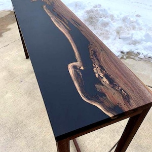 Op de afbeelding: Een lange, rechthoekige houten tafel met een donker, glanzend zwart epoxyhars midden. De randen en poten van de tafel zijn gemaakt van natuurlijk hout, met een rijke bruine nerf. De tafel is ongeveer 183 cm lang.