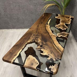 May include: A rectangular wooden table with a live edge design. The table features a clear epoxy resin filling the gaps in the wood, creating a unique and modern look. The table is supported by a black metal frame.