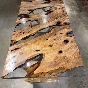 May include: A rectangular wooden table with a clear epoxy resin fill. The wood has a natural, light brown colour with dark brown knots and grain patterns. The table is approximately 183 cm long and 91 cm wide.