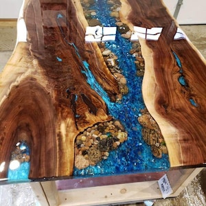 May include: A handcrafted wooden table with a river-like design. The table features a clear blue epoxy resin river with embedded stones, flowing between two sections of rich, dark brown wood. The table's base is made of light-colored wood.
