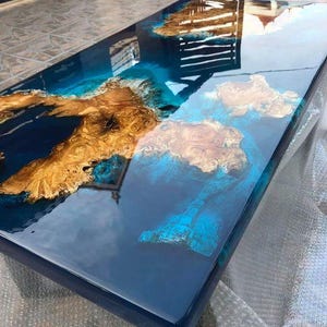 Op de afbeelding: Een rechthoekige epoxyhars tafel met een glanzende afwerking. De tafel heeft een diepblauwe basis met secties van goudkleurig hout en turquoise accenten, wat een uniek, artistiek ontwerp creëert. Het oppervlak reflecteert de omgeving.