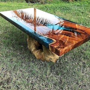 Peut inclure: Une table rectangulaire en bois avec une finition brillante, présentant un motif en résine bleue et transparente. La surface de la table reflète une image de palmier. Le pied est fait d'un tronc d'arbre naturel brun clair.