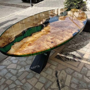 Op de afbeelding: Ovale houten tafel met een glanzende afwerking. Het tafelblad heeft een natuurlijk houtnerfpatroon met een donkergroene epoxyharsrivier. De tafel wordt ondersteund door een zwarte metalen voet. De tafel is ongeveer 150 cm lang.