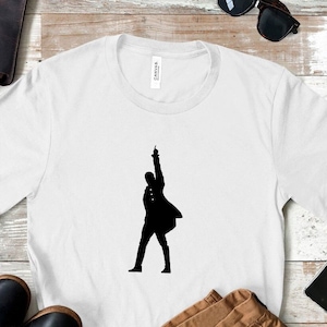 Peut inclure: T-shirt blanc avec une silhouette noire d'une personne levant le bras. Le t-shirt est présenté avec un sac en cuir marron, des chaussures noires, des lunettes de soleil et un pantalon beige sur une surface en bois clair.