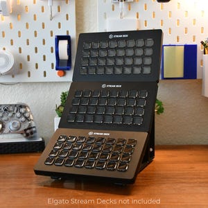 May include: Three black Stream Deck devices with multiple square buttons are displayed on a wooden desk. The devices are stacked, with the top two angled upwards. A clear Xbox controller sits to the left. The text "Elgato Stream Decks not included" is at the bottom.