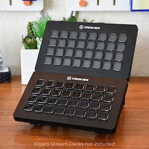 May include: Two black Stream Deck devices with multiple square buttons are displayed on a wooden surface. The Stream Deck logo is visible on each device. The text "Elgato Stream Decks not included" is at the bottom.