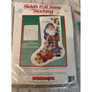 May include: A Sleigh Bell Santa Stocking cross stitch kit by Dimensions. The packaging features a white stocking with a printed design of Santa Claus in a blue coat, toys, and the name Emilee. It includes instructions for a 16 inch finished size.