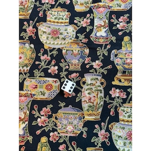 May include: A black fabric with a repeating pattern of ornate vases, teapots, and floral elements in shades of gold, pink, purple, and green. A white die with black dots is placed on the fabric.