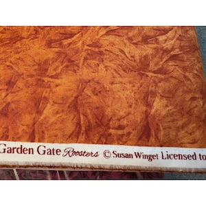 May include: Close-up of a fabric with a textured, mottled pattern in shades of orange and brown. The fabric has a rough, organic appearance. The text "Garden Gate Roosters" and "Susan Winget Licensed to" is printed on a white strip.