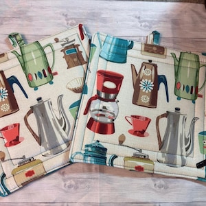 May include: Two square cotton pot holders with a cream background. The pot holders feature a repeating pattern of coffee pots, coffee makers, and cups in shades of brown, green, red, and blue. The edges are trimmed with a blue fabric.