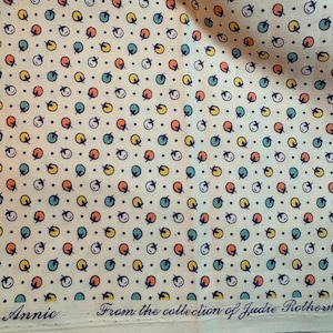 May include: A cream-colored fabric featuring a repeating pattern of small, stylized orange and teal fruit shapes interspersed with tiny black dots. The bottom edge displays the text Annic From the collection of Judie Rothermel.
